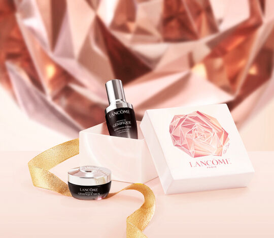 A sleek display featuring Lancome beauty products, including a serum bottle and a cream jar, styled with a decorative ribbon and set against a shimmering rose gold backdrop.