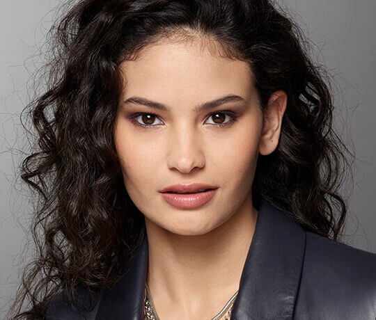 A close-up portrait of a woman with curly dark hair and warm brown eyes, wearing a dark jacket, looking directly at the camera.