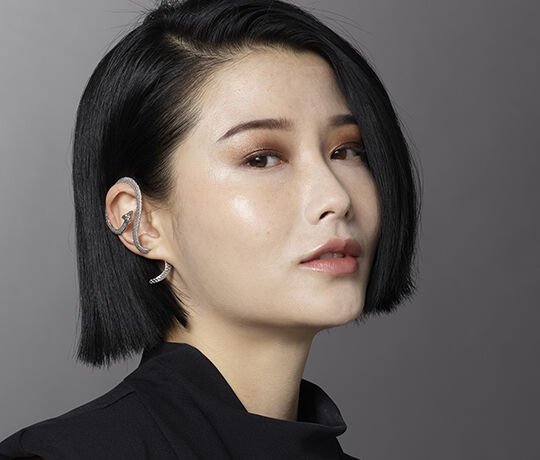 A close-up portrait of a woman with a sleek black bob hairstyle, wearing a stylish earring that resembles a snake. She has soft makeup, with a subtle highlight on her cheek.
