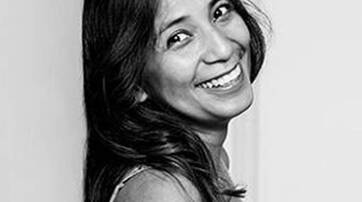 A woman with long, dark hair smiles brightly while leaning against a wall in a black and white photograph.