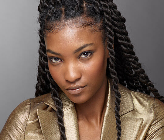 A close-up portrait of a woman with long, twisted hair, wearing a shiny gold blazer, looking confidently at the camera.