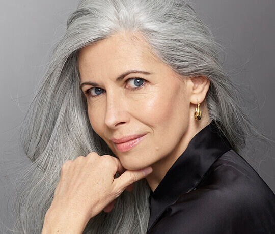 A woman with long, gray hair and striking blue eyes is looking at the camera. She has a soft smile and is resting her chin on her hand, wearing a black top and gold earrings.