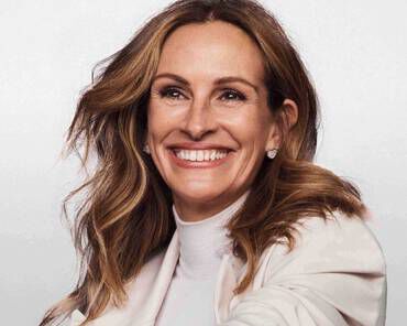 A woman with long, wavy hair is smiling widely, wearing a white top and a light-colored jacket. The background is light and soft, enhancing her joyful expression.