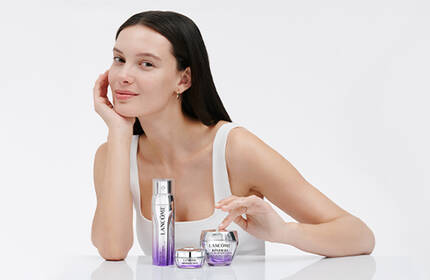 A woman with long dark hair is sitting at a table with skincare products in front of her, smiling gently at the camera. The background is minimalistic and soft in color.