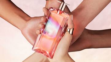 A close-up image of a colorful perfume bottle held in the center by multiple hands, with a soft, dreamy background.