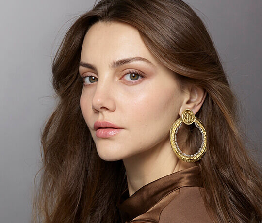 A close-up portrait of a woman with long, wavy hair, wearing large gold hoop earrings and a brown satin top, gazing confidently at the camera.