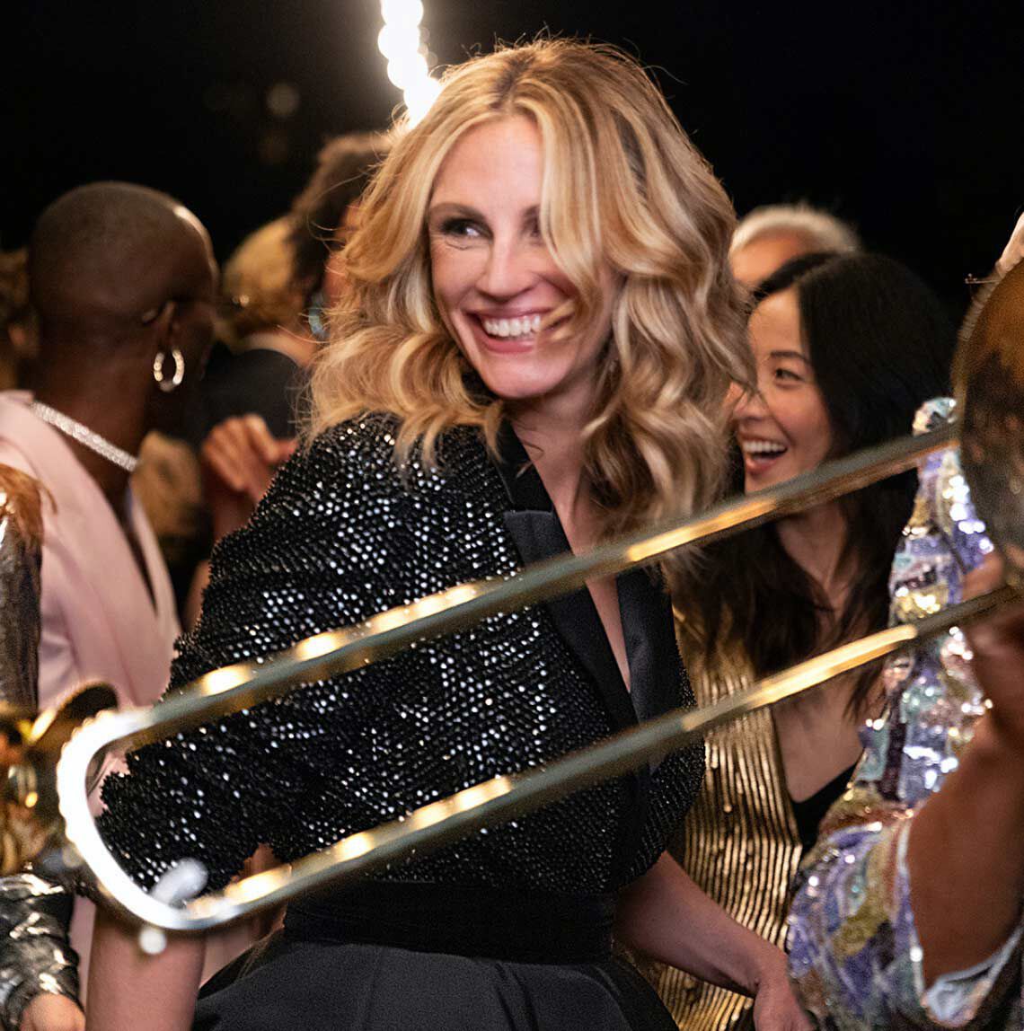 Uśmiechnięta kobieta z falującymi włosami, ubrana w błyszczący żakiet, bawi się na przyjęciu otoczona innymi szczęśliwymi gośćmi.