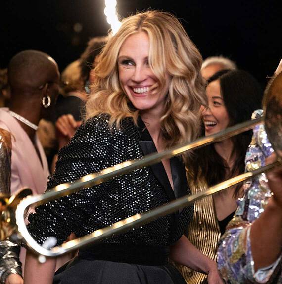 Uśmiechnięta kobieta z falującymi włosami, ubrana w błyszczący żakiet, bawi się na przyjęciu otoczona innymi szczęśliwymi gośćmi.