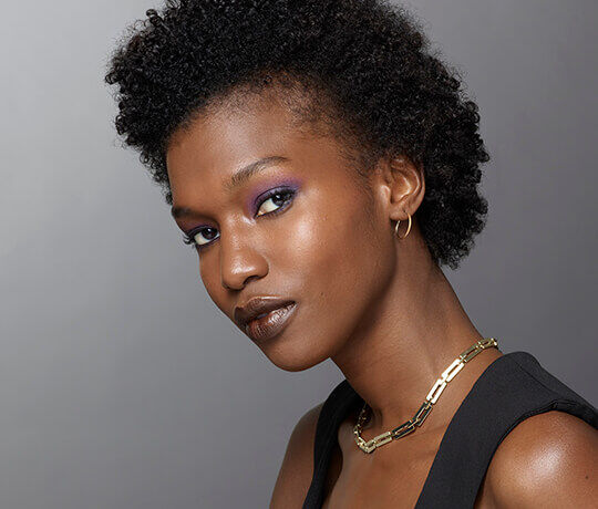 A portrait of a woman with natural curly hair, showcasing vibrant purple eyeshadow, neutral lipstick, and gold jewelry, set against a gray background.