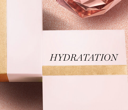 Two elegantly wrapped boxes sit on a textured pink background, featuring the word "HYDRATATION" prominently on one box, with a decorative crystal in the background.
