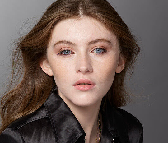 A close-up portrait of a young woman with long, flowing hair, striking blue eyes, and a natural makeup look. She has a soft expression and is wearing a dark, stylish jacket.