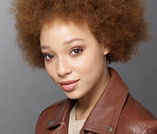 A young person with curly brown hair and freckles is smiling softly at the camera, wearing a brown leather jacket over a light top. The background is a muted gray.