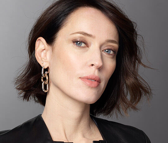 A close-up portrait of a woman with short, dark hair and striking blue eyes, wearing a black leather top and a large chain-link earring. Her makeup is elegant, featuring a natural look.