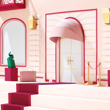 A whimsical display featuring a pastel pink building facade with a large arched door, adorned with a soft pink awning. Colorful geometric shapes and boxes are placed on the steps, and elegant ropes and stanchions create an inviting entrance.