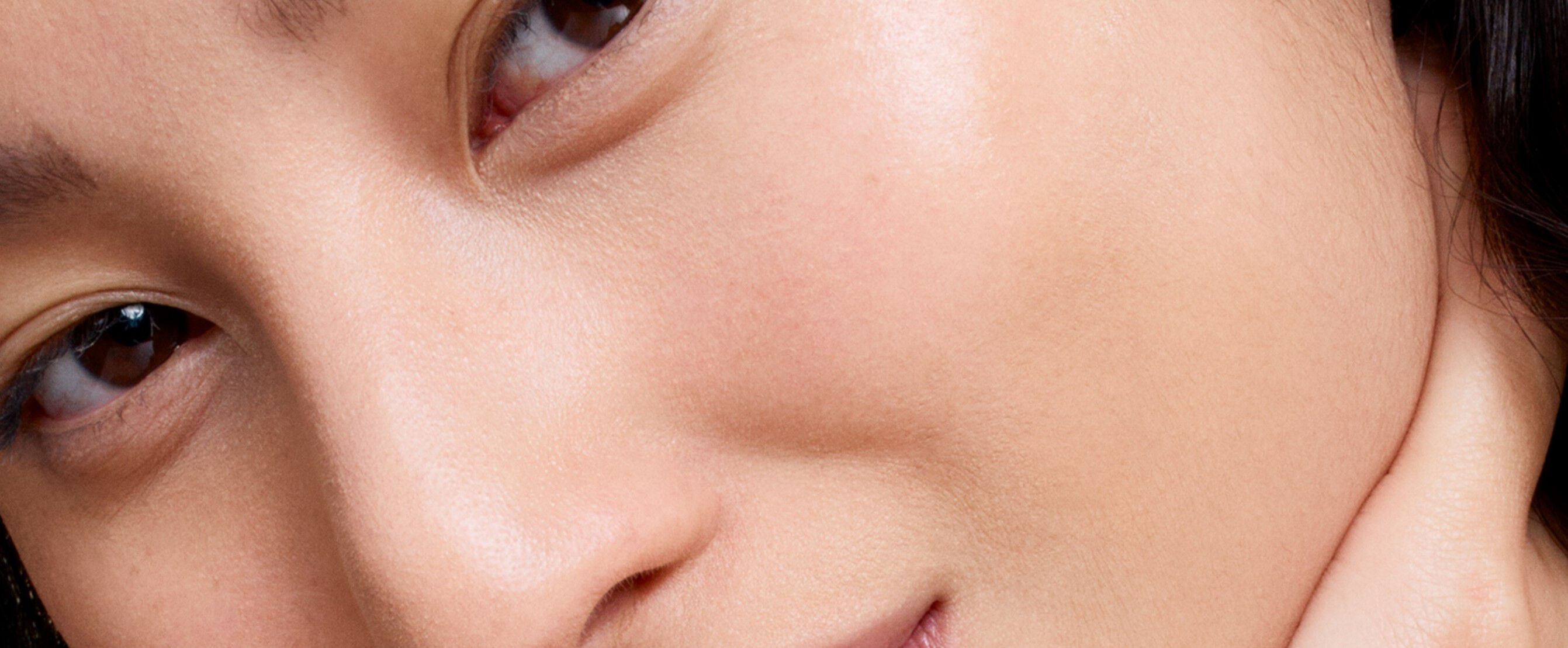 Zbliżenie na twarz osoby z delikatnym uśmiechem, widoczne oczy i gładka skora. Tło czarne.