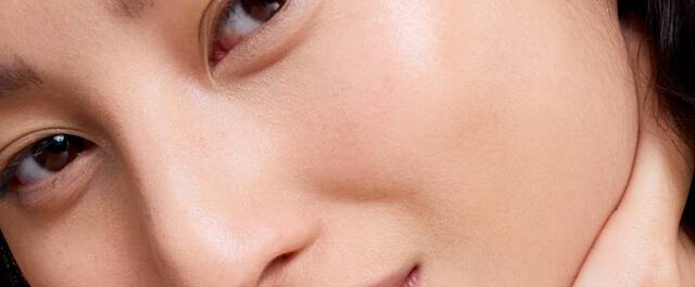 Zbliżenie na twarz osoby z delikatnym uśmiechem, widoczne oczy i gładka skora. Tło czarne.