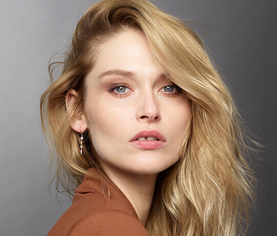 Close-up portrait of a woman with long, wavy blonde hair and blue eyes, wearing subtle makeup and silver earrings, against a neutral background.