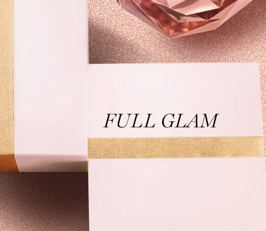 A close-up of two elegant gift boxes in soft pink, one adorned with a gold stripe, resting on a sparkling surface with a crystal decoration in the background. The text "FULL GLAM" is prominently displayed on the box.