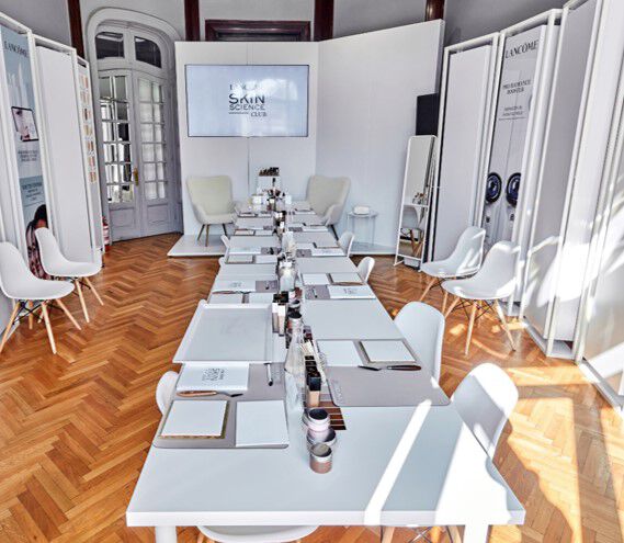 Imaginea prezintă o incăpere elegantă cu mobilier modern, in care se află o masă lungă, pregătită pentru un eveniment, cu scaune albe și lumina soarelui care pătrunde prin feronerie. Pe pereți sunt panouri cu brandul "Lancome".