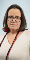 A woman with dark hair and glasses, wearing a light-colored top and a red and black beaded necklace.