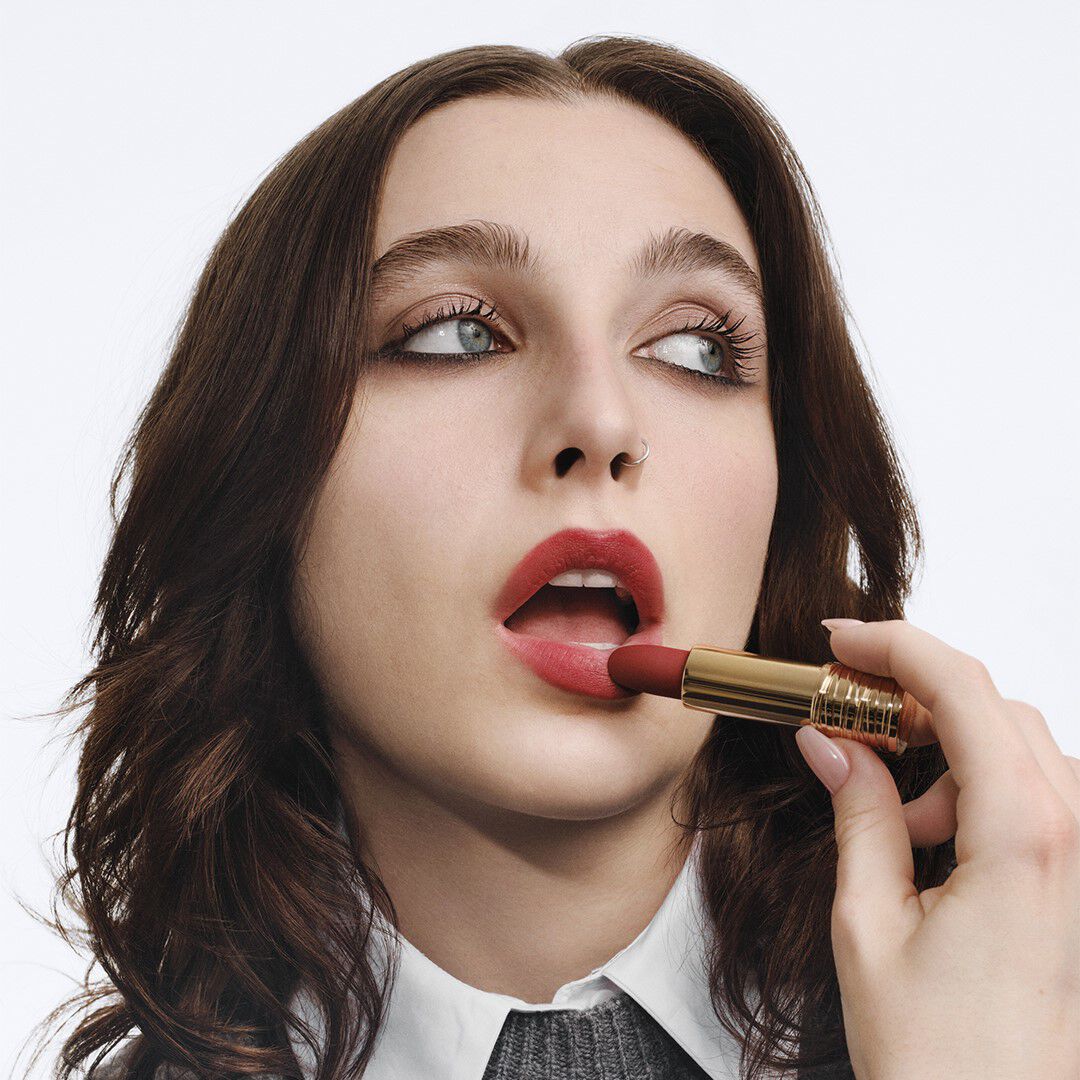 Młoda kobieta nakłada czerwony pomadkę, patrząc w gorę, z długimi, falującymi włosami i delikatnym makijażem.