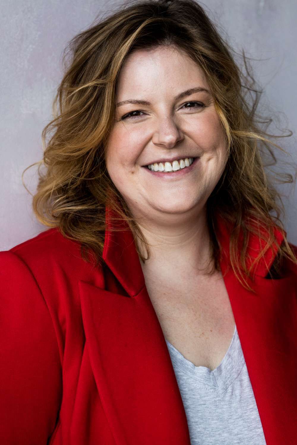 A smiling woman with wavy brown hair, wearing a red blazer over a grey shirt.