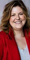 A smiling woman with wavy brown hair, wearing a red blazer over a grey shirt.