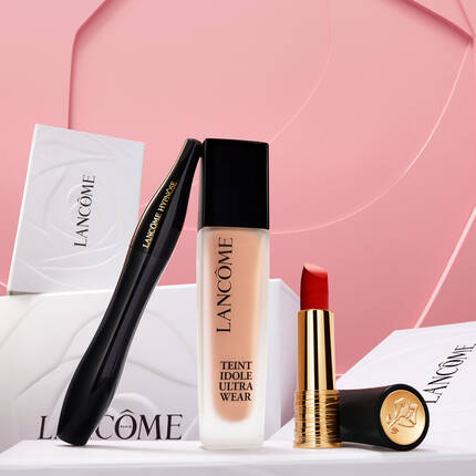 Zestaw kosmetykow marki Lancome, w tym tusz do rzęs, podkład Teint Idole Ultra Wear oraz pomadka w soczystym odcieniu, na tle rożowej faktury.