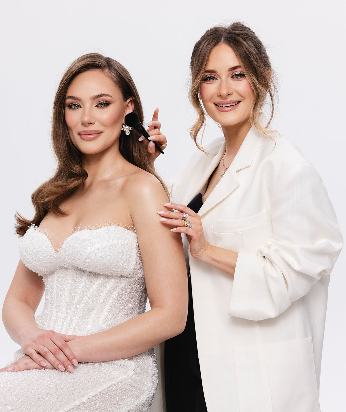 Dwie kobiety na białym tle. Jedna kobieta, ubrana w białą, cekinową suknię ślubną bez ramiączek, siedzi na krześle. Druga kobieta - makijażystka, w białej marynarce, stoi za nią, delikatnie dotykając jej twarzy pędzlem do makijażu. Obie uśmiechają się.