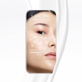 Zdjęcie przedstawia smartfon z aplikacją Lancome e-SKIN EXPERT, na ktorej widoczny jest zbliżony portret osoby oraz wskaźnik promienistości na poziomie 65%. W tle znajduje się biały, falisty wzor.