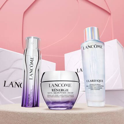 Trzy produkty kosmetyczne marki Lancome: serum, krem przeciwstarzeniowy i esencja rozjaśniająca, ustawione na rożowym tle.